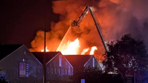 Orange flames and a plume of smoke coming from a warehouse at an industrial site. A fire engine aerial ladder is being used to put out the fire.