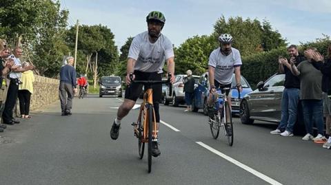 Two men wearing white t-shirts on bikes as they get clapped on at the finishing line on a road in Ballaugh.