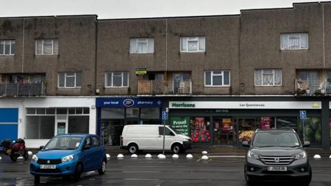 Three shops in a 1960s-looking terrace in Les Quennevais. They include an empty shop unit, a small Morrisons supermarket and a Boots pharmacy.