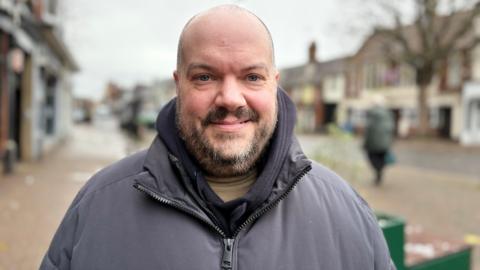 Chris Leaman with very short hair and dark beard smiling at the camera while wearing a grey anorak in Leighton Buzzard High Street with shops in the background and a shoppers in a green coat to the right.