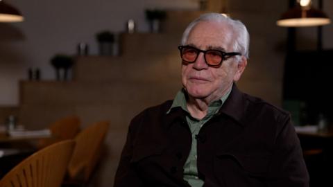 Brian Cox sits in a dark room, wearing dark tinted glasses, a green shirt and dark jacket