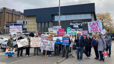 A group of men and women, outside a large building, holding placards with messages campaigning for the reopening of the leisure centre. The closed building can be seen in the background.  