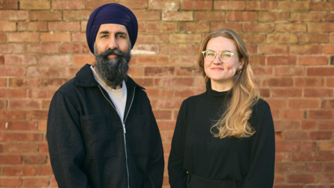A man wearing a turban and jacket and a woman wearing glasses and a dark top stand side by side in front of a brick wall.
