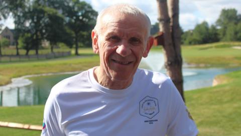 Peter Reid stands in a park in front of a small lake on a sunny day. Reid, 70, has silver short hair and is leaning on a metal gate. He is wearing a t-shirt which says 'The Sir Bobby Robson Foundation'.