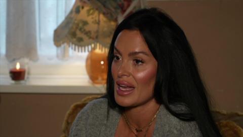 A woman with black hair and wearing a grey shirt and gold necklace. She is sitting in a living room.