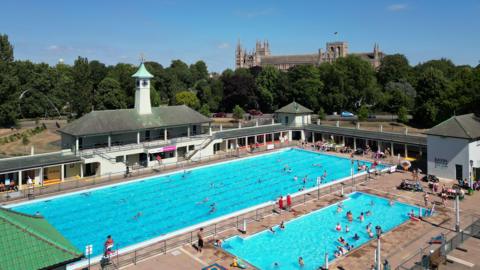 Peterborough Lido, which has two swimming pools - one bigger than the other - with people swimming and lifeguards sitting to the site. The pools are surrounded by white walls with a grey roof and are overlooked by a clock tower with a light blue top. Peterborough's cathedral is obscured by trees.