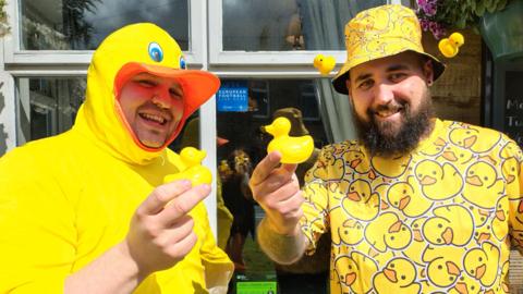 Daniel and Curtis are both smiling at the camera while holding a single rubber duck each. Daniel is dressed in a yellow rubber duck costume and Curtis is wearing a hat and t-shirt which are covered with pictures of yellow rubber ducks.