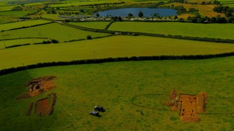 An aerial view of the excavation sites. Two sections of a green fields have been dug up, revealing the soil underneath.