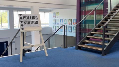 The photo shows an indoors sign which says polling station with arrows pointing down some stairs. The sign is on some blue carpet. There are windows in the background, and artwork is on the wall.