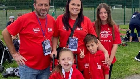 Young girl Elodie, who is sat in a wheelchair and has a tube going into her nose, poses with her parents and two siblings. The family are wearing matching red t-shirts and hoodies for the British Transplant Games.