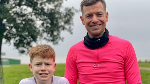 Ralphy and Stuart Raffle in Harwich. Ralphie is aged 10 and has short light reddish hair and is wearing a white T-shirt. On his left is Stuart who has short grey hair and is wearing a pink running top and black neck tube. Behind them is a wide path with grass and a tree.