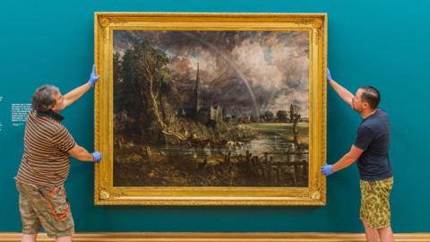 The large oil painting, Constable's Salisbury Cathedral from the Meadows, is being hung by two staff at the Tate Britain - its ornate gold frame contrasting against the teal coloured wall. In the foreground of the painting there is a horse drawn cart through the water meadows. Behind is Salisbury Cathedral and spire and in the distance some buildings, including Leaden Hall. A rainbow arcs through the cloudy sky towards the hall. 