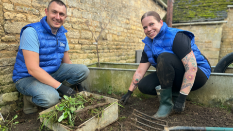 Graeme and Katherine, both wearing blue gilets, kneeling down planting trees 