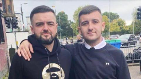 Two young men stand in an urban street with their arms round each other's shoulders. The one on the left has dark hair, a beard and is wearing a black hoodie. The one on the right has short dark hair, and is wearing a blue jumper over a white shirt.