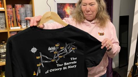 Eva White holding one of the Tar Barrels merchandise t-shirts. She has long brown hair and is wearing a pink hoodie.