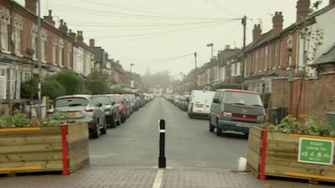 Street in Kings Heath, Birmingham, a bollard in the middle with two wooden boxes each side contains plants. Cars are parked on either side of the road next to houses.