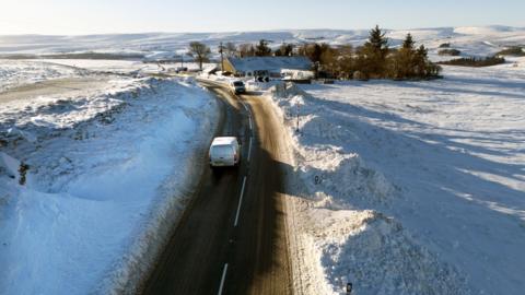 A van drives along a road where fields on either side have been covered by snow. A farmhouse is located to the right of the road.