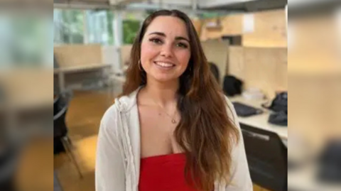 The image shows a girl with long brown hair smiling; she is also wearing a red top and white cardigan. Behind her desks and chairs can be seen.