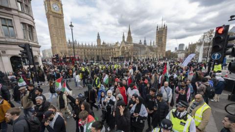 Large march in central London with the Houses of Parliament behind a crowd of marching protesters. Some are holding Palestinian flags