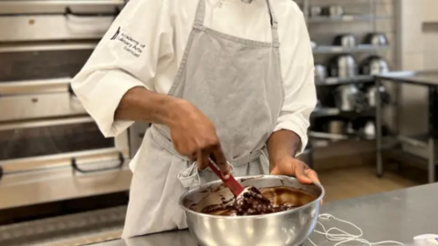 A man folding a chocolate mixture in a cooking bowl in a steel kitchen. He is wearing an apron and apprentice white shirt.