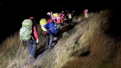 A mountain rescue team wearing helmets and large bags carry a stretcher over uneven terrain in the dark. 