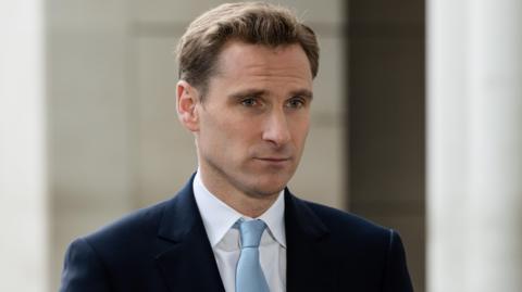 Shadow home secretary Chris Philp, pictured in a dark blue suit and light blue tie. He has a side parting in mousy brown hair and is looking directly forward.