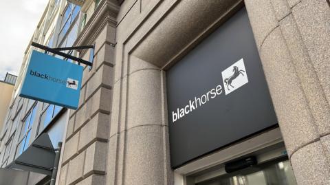 A black sign with white writing which says 'black horse', next to an image of a black horse on a white background. The sign is positioned above a door, and is surrounded by granite brickwork. To the left of the image is a light blue sign which also says "black horse".