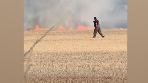 A fire man walking across a field with fire in the background 