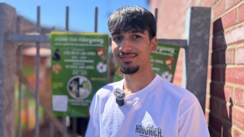 A teenager with dark hair and facial hair, wearing a white tshirt. He is stood in front of a silver gate and brick wall, with his green poster for the "alive the alleyway" mission next to him. The poster is calling for volunteers to help out.