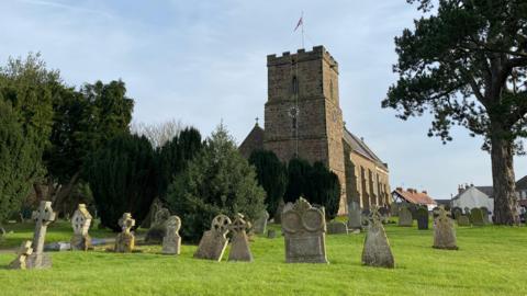 A church is visible in the background, with a newly refurbished clock. In the foreground is a graveyard with oak coloured graves. 