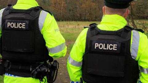 Two police officers standing with their back to the camera wearing a hi-vis police jacket and a black police bullet proof vest, they are also wearing police hats.