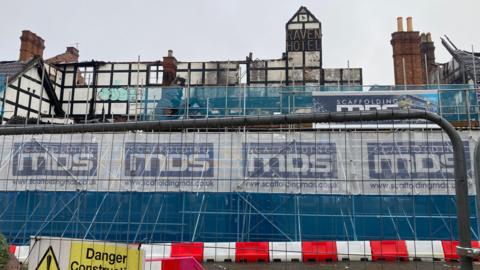 A large black and white Tudor building, with sections of the site discoloured from a fire and holes in the wall. 