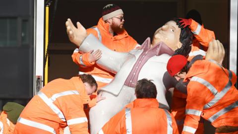 Five men in high viz orange jackets, carrying one of Beryl Cooks sculptures out of a van. The sculpture is of 'Dancing Tom'.