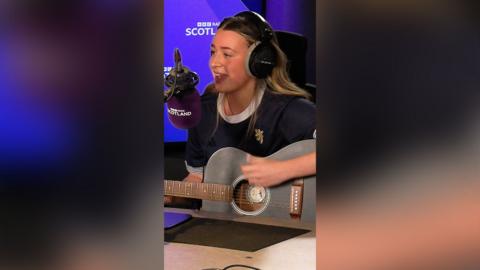 Farrah Robb, a musician with long brown/blonde hair in the BBC Radio studio. She is wearing a blue Scotland football top and is playing a dark grey guitar.