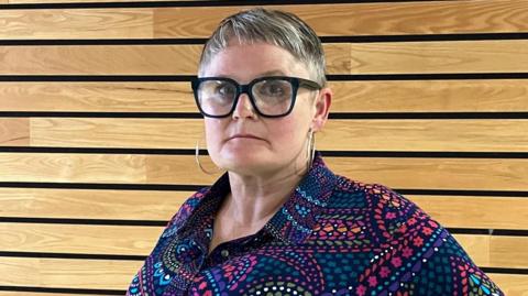 Mae gan Joe William sbectol gyda ffram ddu, gwallt byr ac mae'n gwisgio crys lliwgar. Mae'n edrych mewn i gyfeiriad y camera.