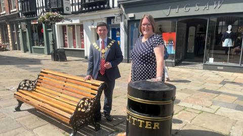 A man with dark hair in a blue suit and red tie and mayoral chain standing behind a wooden slatted bench with black metal sides. Next to him, a woman with brown hair and a black dress with white spots is standing behind a black metal litter bin with the word litter in gold writing. They are on a paved street with shop fronts in the background.
