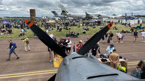 A plane's front propellers dominates the frame. Then a runway with many people standing around, and more aircrafts in the background.