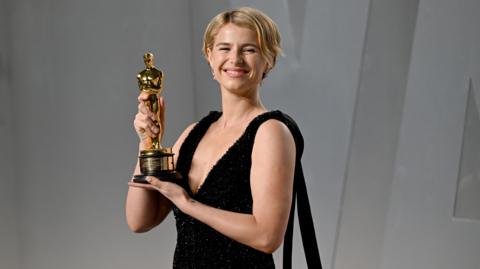 Jessie Buckley with short blonde hair holds up a gold Oscar award with both hands while wearing a black, sleeveless dress
