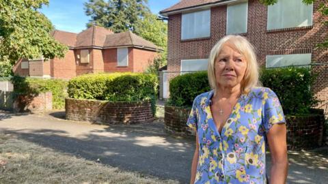 A woman with blonde hair and a flowery blue dress stands outside two large houses with hedges and a driveway. The windows of both houses are bordered up and the gardens surrounded by metal fencing.