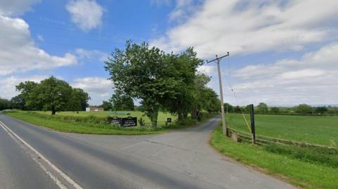 A tree-lined street off the A6108 leads down to the cricket club. Lush green fields are to the left and right of road and a sign that reads 'West Tanfield Cricket Club' can be found to the left, near the entrance to the side road. 