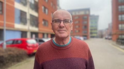 An elderly man with close cropped hair and glasses stands in front a complex of red-brick flats