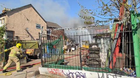 A fire officer pulls a hose across an industrial yard containing tyres, trailers and scrap materials. Plumes of smoke and a jet of water are at the back of the yard.