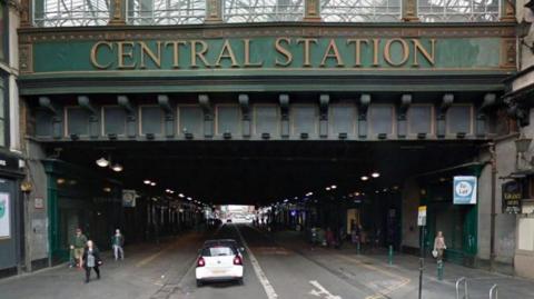 A large railway bridge, with signage saying CENTRAL STATION, overlooking a road and some shops. 