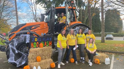 Five women wearing yellow t-shirts with "Zoe's Place' written on. They are standing or sitting on haystacks and there is also an orange tractor with net shaped like cobwebs on it and the word "Zoe's Place Boo Bash" in blue, orange and green writing.
