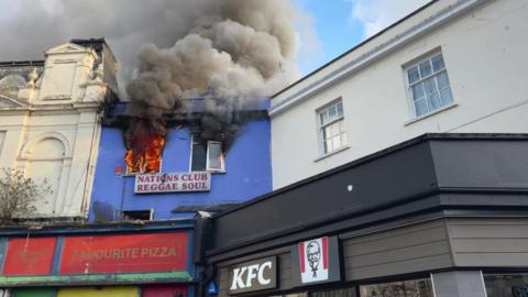 Smoke coming from a window which has flames coming out of it, there is a sign underneath which reads Nations Club Reggae Soul and next to it underneath a sign which reads KFC. 