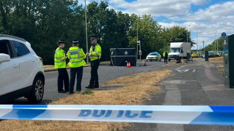 An incident behind a police tape cordon, with uniformed police officers talking and a black privacy screen erected in the middle of a road