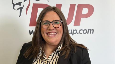 Shows a woman with glasses and smiling in front of a DUP banner, which says DUP and has a lion on the motif. 