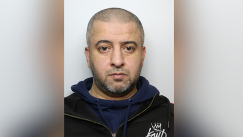 A police custody shot of Fazal Mateen, who has a shaved head and a short goatee beard. He is wearing a black zipper with a blue hoody.