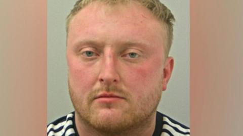 Zak Honeyman's police mugshot, showing a white man with rosy cheeks and pale eyes, short blond hair and a short beard. He is wearing a black and white striped top.