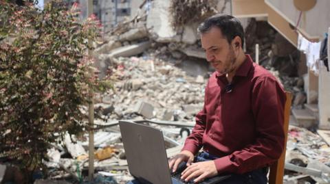 A Palestinian man works on a laptop on a chair outside, surrounded by rubble, in Gaza 
